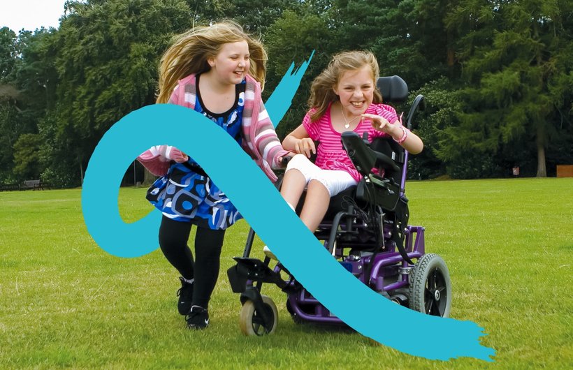 A young wheelchair user plays in her powered chair with her sister running alongside her