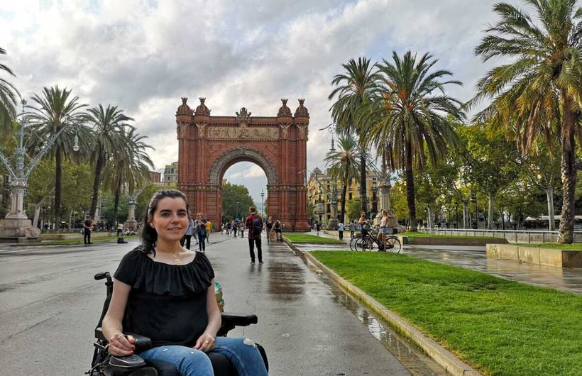 Emma Muldoon in a wheelchair on a palm tree lined road with a large stone arch at the end.