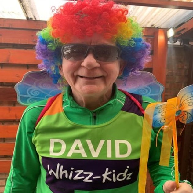 David is wearing a green Whizz Kidz running vest, with a colourful wig, wings and cool magic wand