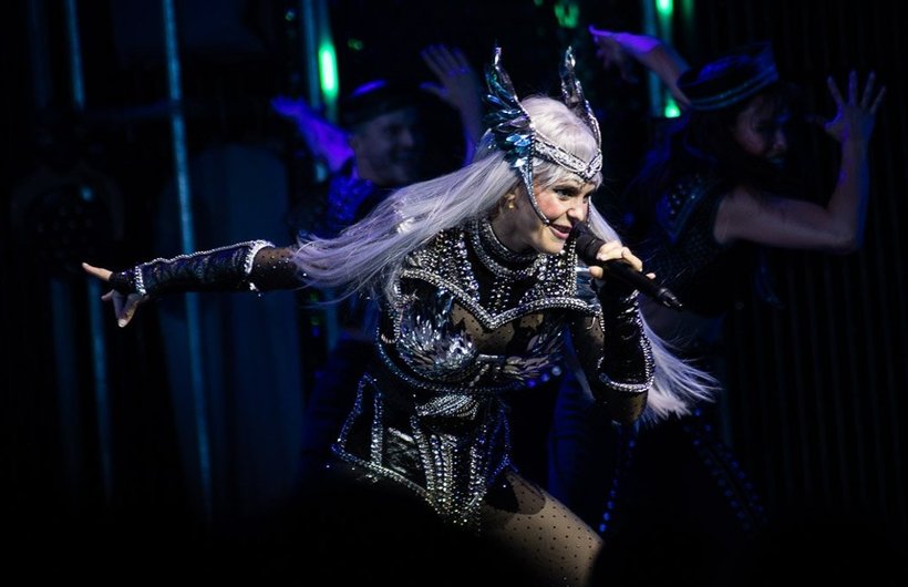 A performer on stage. They have long silver hair, a winged head dress and a black leather costume. They are singing into a microphone.