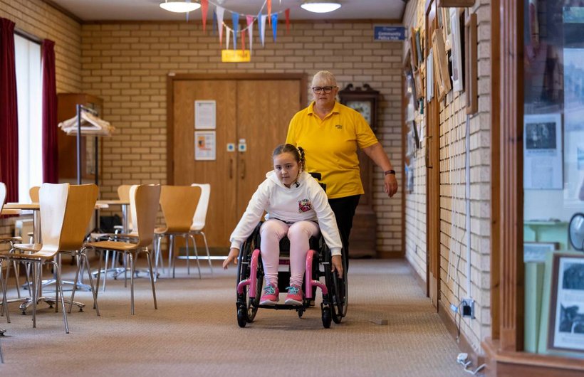 A young person propelling her wheelchair towards the camera in a clinic setting. Sarah Wallace is behind her, wearing a yellow top.