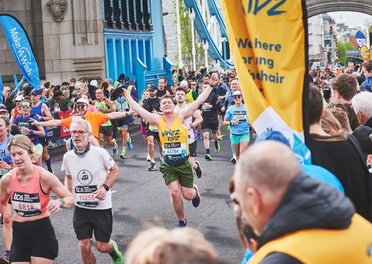A runner in a yello Whizz Kidz running vest holds his arms in the air as he approaches a Whizz Kidz cheer point