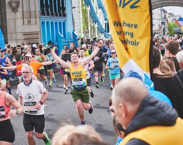 A runner in a yello Whizz Kidz running vest holds his arms in the air as he approaches a Whizz Kidz cheer point