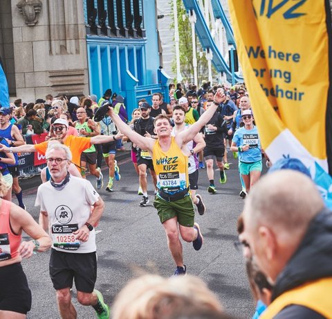 A runner in a yello Whizz Kidz running vest holds his arms in the air as he approaches a Whizz Kidz cheer point