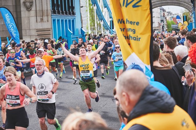 A runner in a yello Whizz Kidz running vest holds his arms in the air as he approaches a Whizz Kidz cheer point