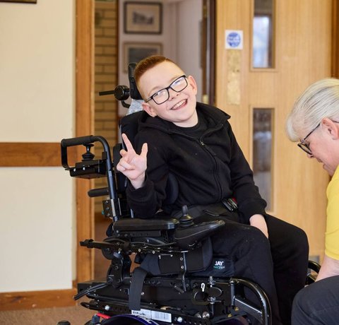 Logan smiling at the camera whilst Sarah, a Whizz Kidz clinician adjusts their wheelchair