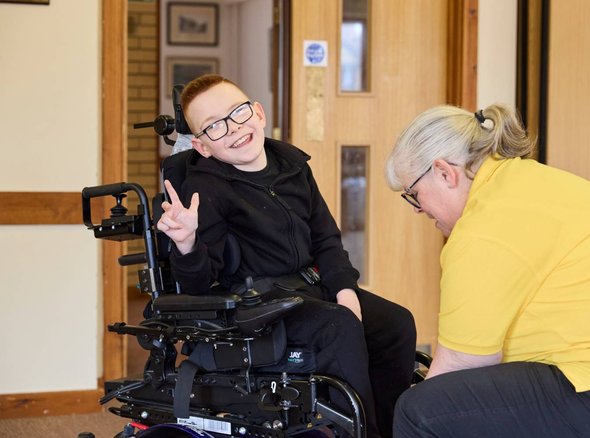 Logan smiling at the camera whilst Sarah, a Whizz Kidz clinician adjusts their wheelchair