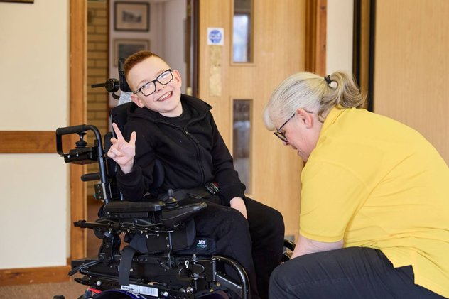 Logan smiling at the camera whilst Sarah, a Whizz Kidz clinician adjusts their wheelchair