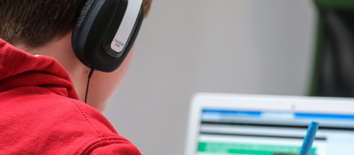 A young person with earphones on holds a pen whilst looking at a laptop computer screen