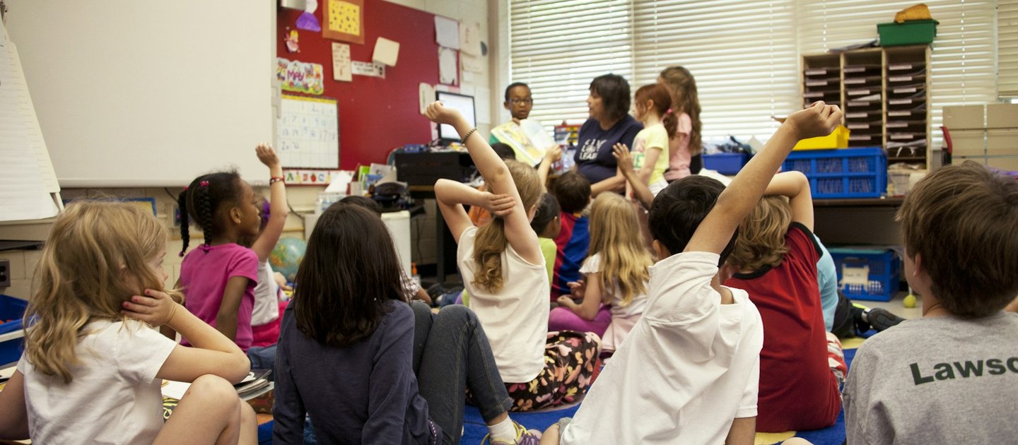 A group of school children sit on the floor. Some have their hands up as they listen to the teacher.