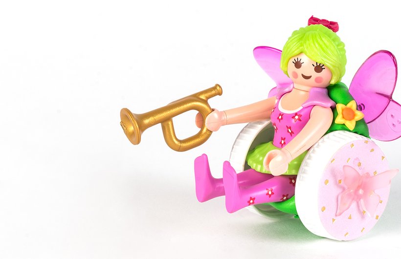 A Playmobile fairy figure sitting in a brightly coloured wheelchair holding a golden trumpet.