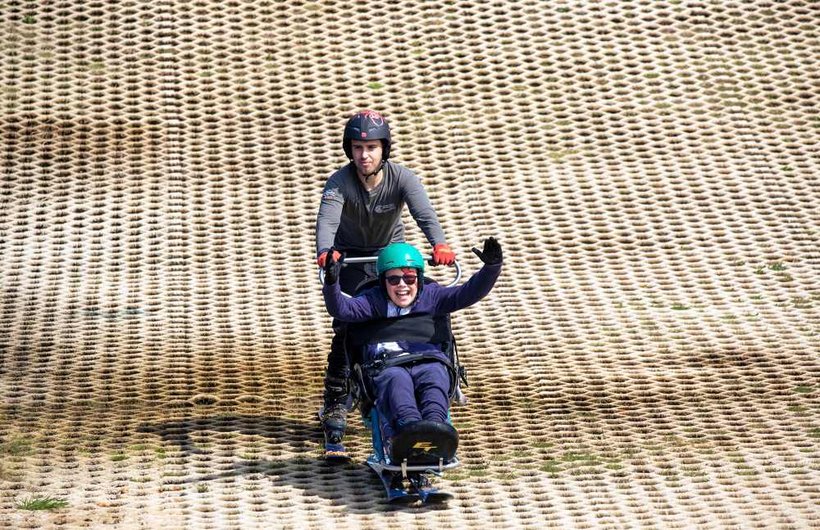 A young wheelchair user is pushed down a dry ski slope in adapted skis
