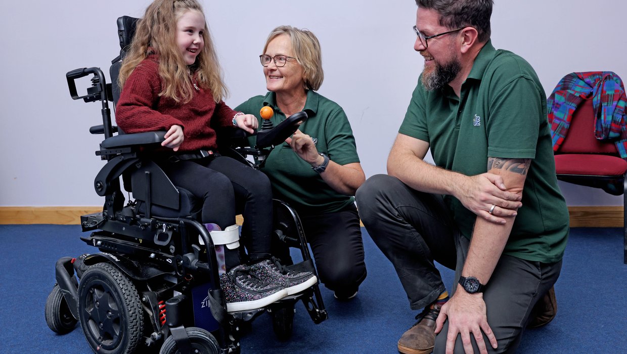 Margot, a six year old girl using a powered wheelchair, smiles at Whizz Kidz staff at a clinic