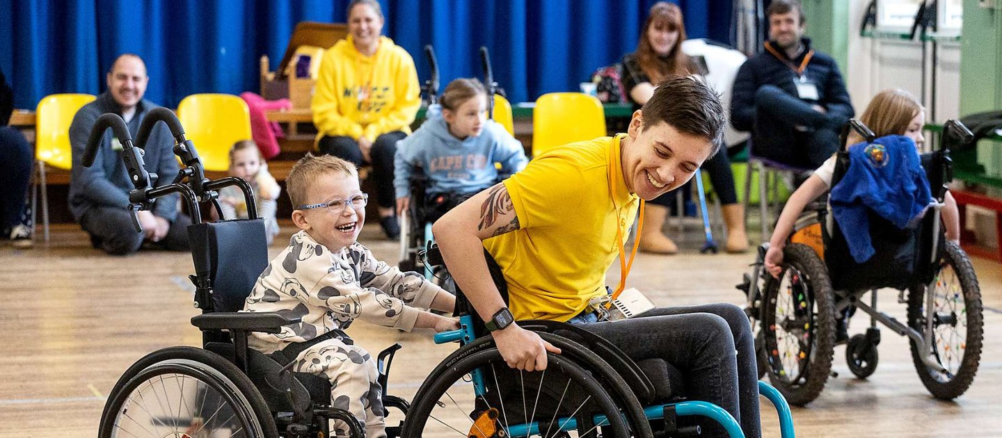 Nix, our skills trainer pulls along a smiling young wheelchair user for a ride. People look on smiling in the background of the sports hall