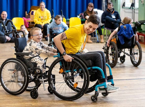 Nix, our skills trainer pulls along a smiling young wheelchair user for a ride. People look on smiling in the background of the sports hall
