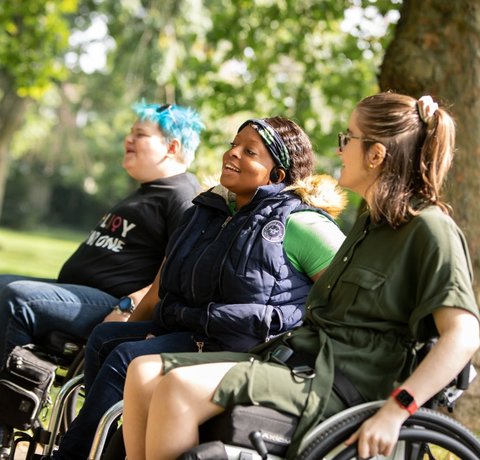 Three young wheelchair users sit laughing and smiling in a sunny park
