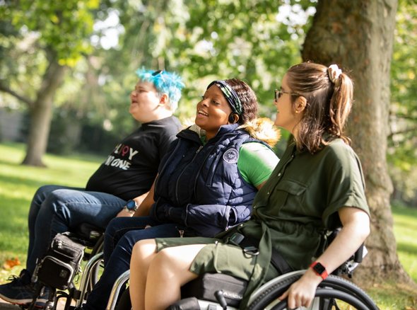 Three young wheelchair users sit laughing and smiling in a sunny park