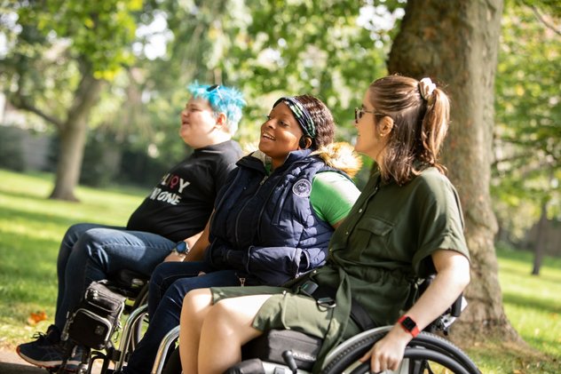 Three young wheelchair users sit laughing and smiling in a sunny park