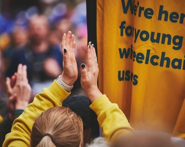 People clapping on the Whizz Kidz cheer point next to a Whizz Kidz flag which reads "we're here for young wheelchair users"