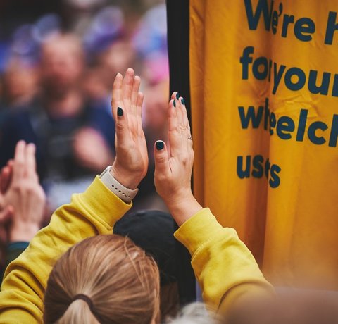 People clapping on the Whizz Kidz cheer point next to a Whizz Kidz flag which reads "we're here for young wheelchair users"