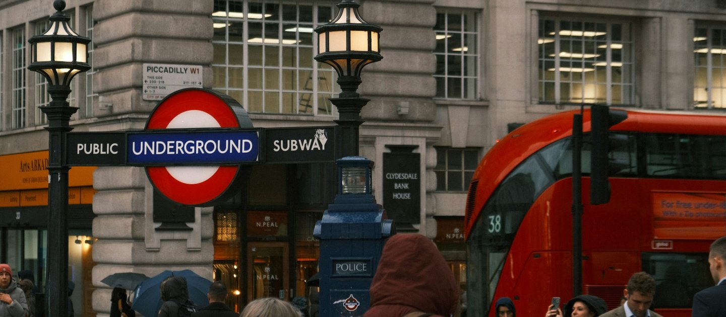 A London Underground sign with a red bus going past it and lots of people in the foreground