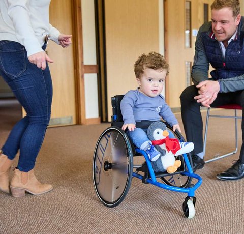 Jake in his wheelchair looking at the camera. He has his toy penguin on the footplate. He is wearing blue Adidas trainers.