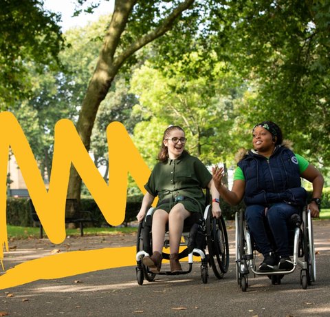 Two young wheelchair users laughing in a park area surrounded by trees
