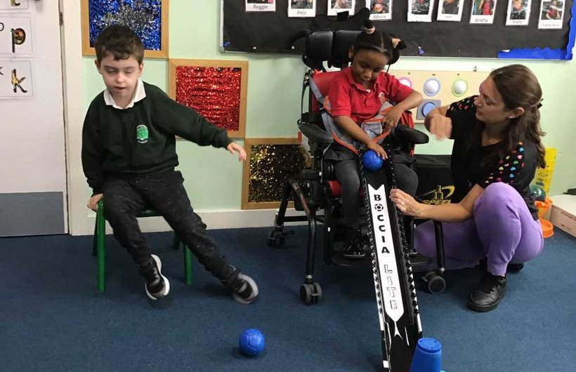 A young wheelchair user plays boccia with an adult. There is another child sat on a chair next to them