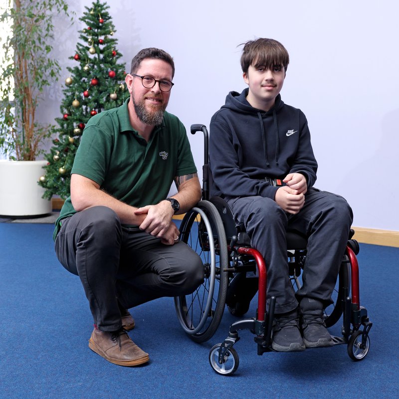 Archie, a teenager with dark hair, sits in his wheelchair next to a Whizz Kidz technician