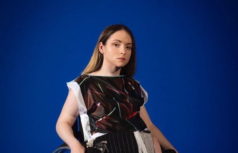 Caitlyn Fulton in a wheelchair, wearing a dark red top with white vertical edges, and thin light blue rectangles at irregular angles. Her skirt is black with white pinstripes and she is wearing black shoes. She has a neutral expression and her right hand