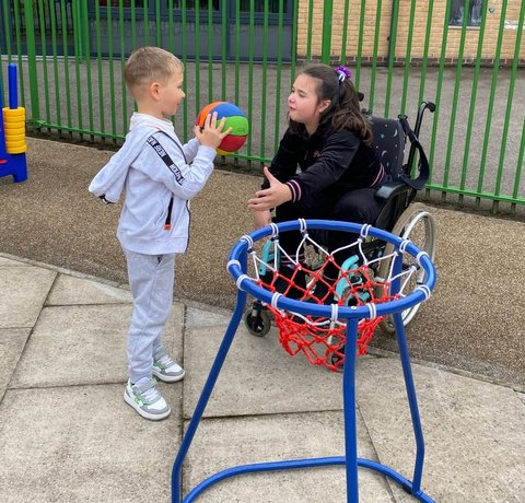 Two children play with a ball next to a low basketball hoop. One child is wearing a grey tracksuit and the other wearing a black top and is sitting in a black wheelchair.