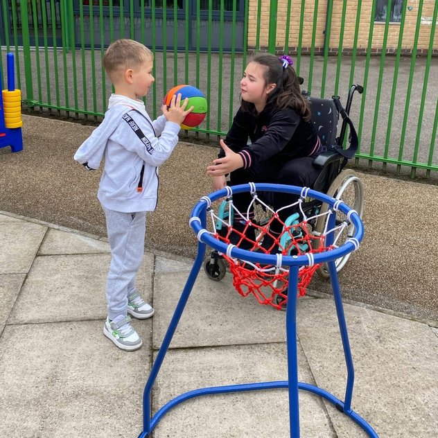 Two children play with a ball next to a low basketball hoop. One child is wearing a grey tracksuit and the other wearing a black top and is sitting in a black wheelchair.