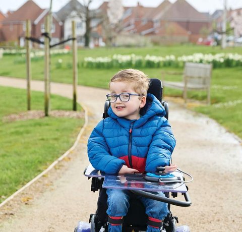 Ethan is wheeling forward in his powered chair. He is wearing a blue puffer jacket and going down a pathway through a park