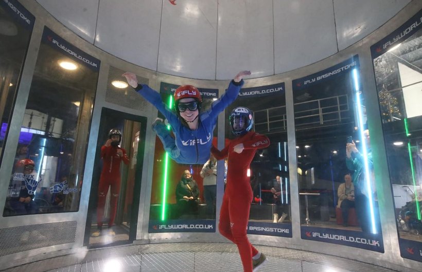 Rebecca indoor skydiving. She is hovering in the air, smiling, wearing a blue flying suit. Her arms and legs are out stretched.