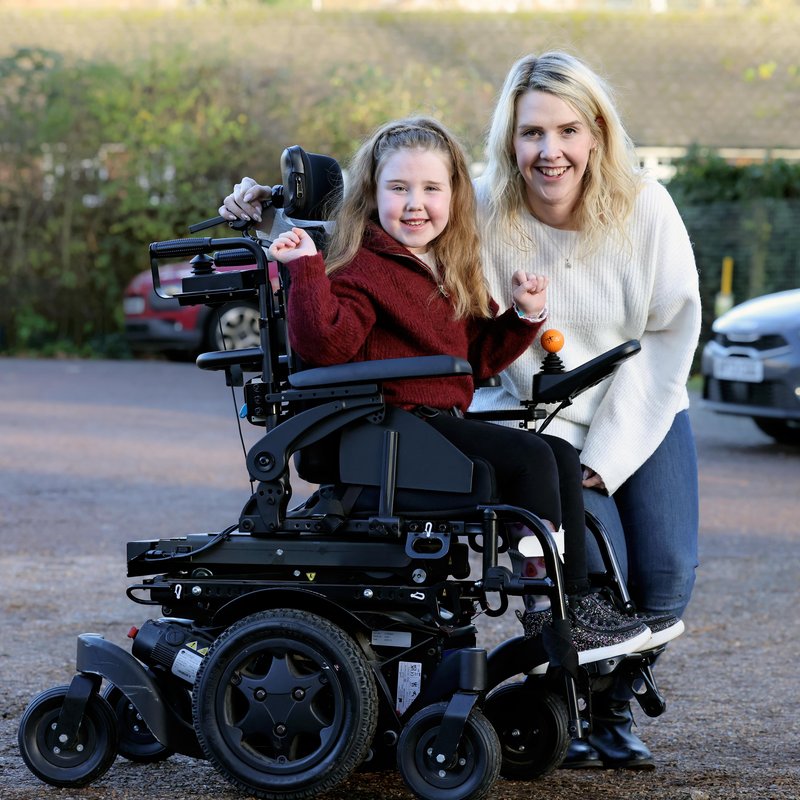 Margot, a six year old girl smiles in her powered wheelchair alongside her Mum in an outdoor space
