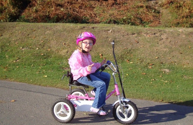 Penny when she was a child, riding a pink trike. She is wearing a pink polka dot jacket and pink helmet. She is outside, looking at the camera and smiling.