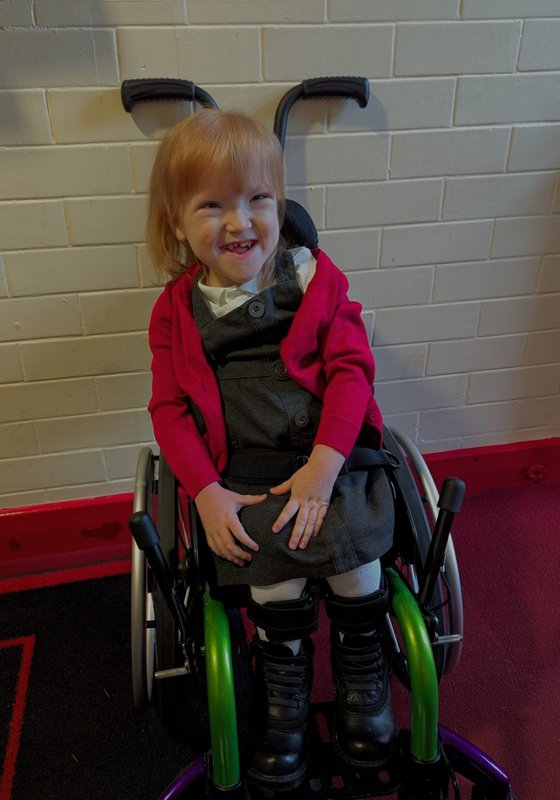 Charlie sits in her green and purple wheelchair, wearing her school uniform.