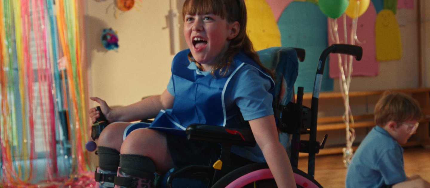Amber, with a big smile on her face, wheels around at an after school club. There are balloons and bright colours in the background.