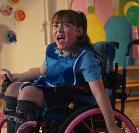 Amber, with a big smile on her face, wheels around at an after school club. There are balloons and bright colours in the background.
