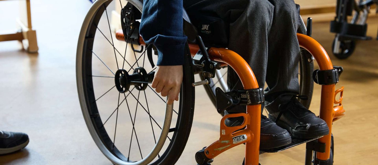 A close up of a smart looking wheelchair with an orange frame. A young wheelchair user has their right hand on one of the wheels.