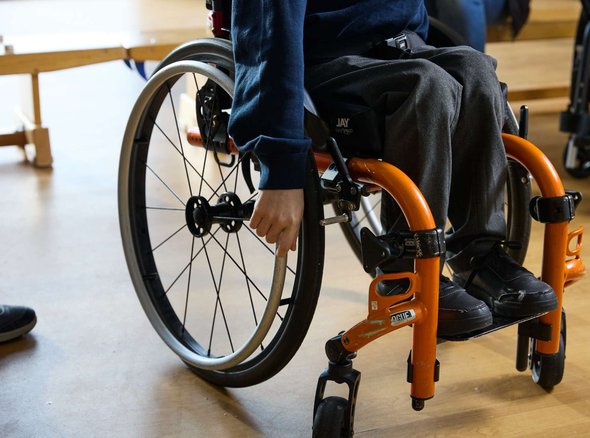 A close up of a smart looking wheelchair with an orange frame. A young wheelchair user has their right hand on one of the wheels.
