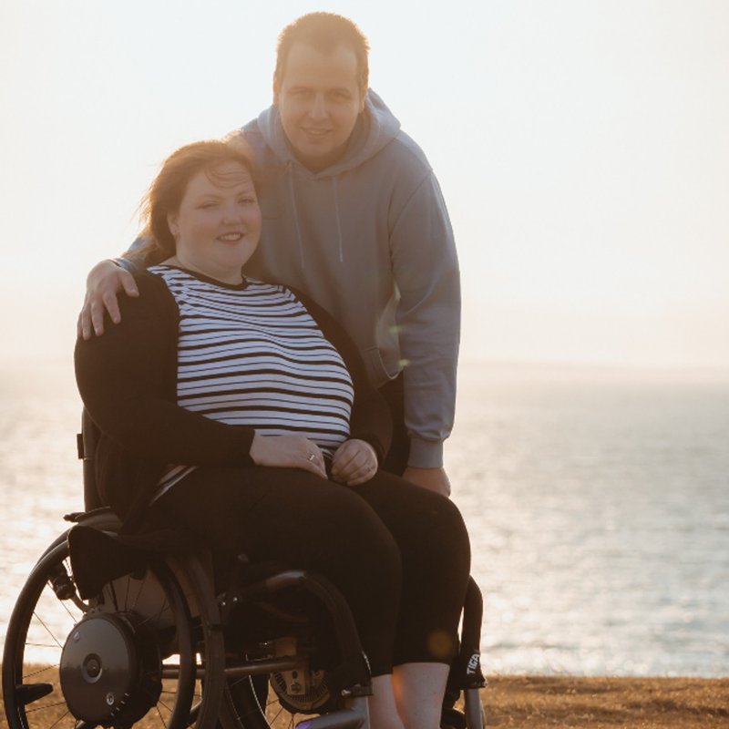 Sophie in her wheelchair with Charlie behind her, standing. They are in front of the sea.