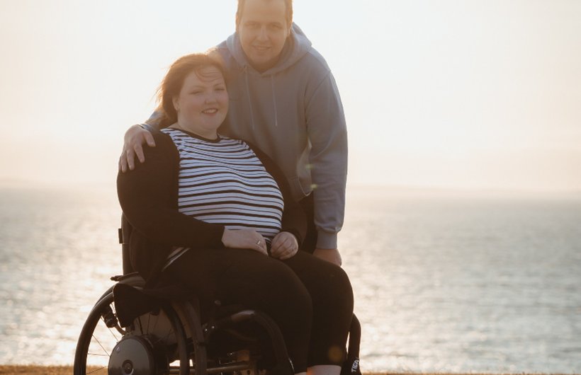 Sophie in her wheelchair with Charlie behind her, standing. They are in front of the sea.