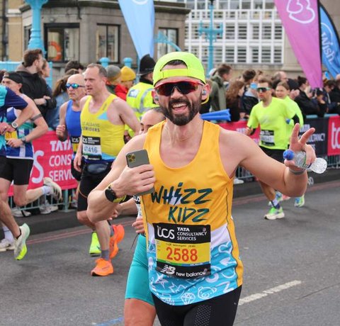 A cool looking runner wearing a yellow Whizz Kidz running vest, with sunglasses and a yellow hat gives the camera a peace sign as he runs past