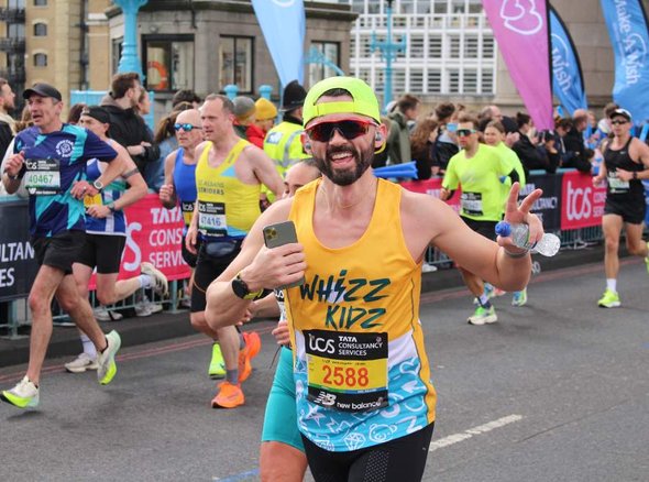A cool looking runner wearing a yellow Whizz Kidz running vest, with sunglasses and a yellow hat gives the camera a peace sign as he runs past