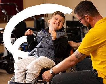 A young wheelchair user smiles as a Whizz Kidz Wheelchair Technician makes adjustments to his chair