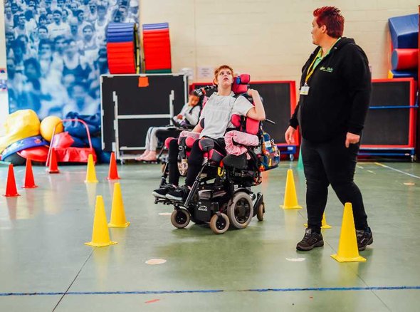 A Whizz Kidz clinician helps a young wheelchair user navigate some small cones in her powered chair