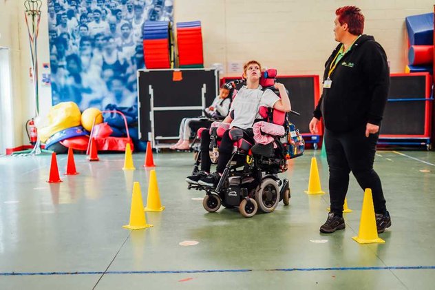 A Whizz Kidz clinician helps a young wheelchair user navigate some small cones in her powered chair