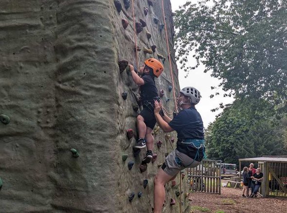 A young wheelchair user climbs up a climbing wall supported by an adult