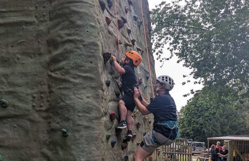 A young wheelchair user climbs up a climbing wall supported by an adult